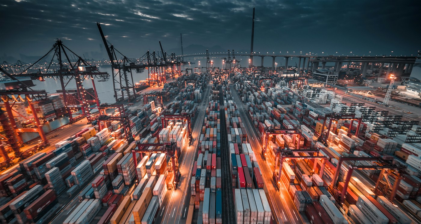 Aerial view of a shipping port with stacked containers and cranes, showing the scale of global freight operations